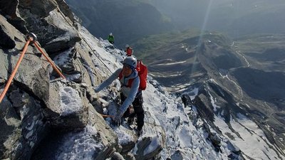 People with helmets and climbing gear are climbing up a secured mountain path; snowy alpine landscape.