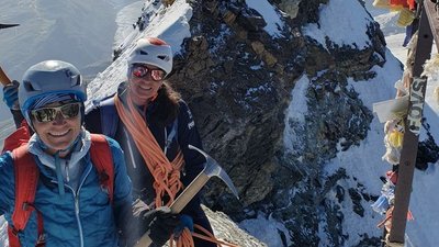 Two people with helmets and ice axes mountaineering on a snow-covered peak in the Alps.