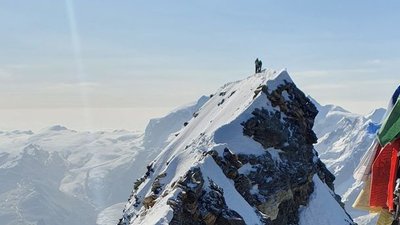 Mountaineer with helmet on snow-covered summit, surrounded by mountains; colorful flags in the foreground.