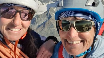 Two people with helmets and sunglasses in front of snow-covered mountains, one of them carrying a rope.