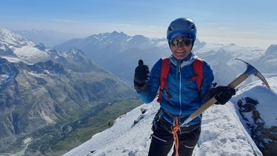 Person with helmet and ice pick on snow-covered mountain peak, with the Alps panorama in the background.