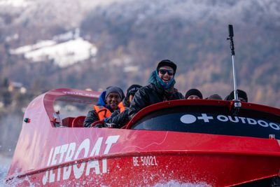 People on a red jet boat wearing jackets and hats, cruising on water with snowy mountains in the background.