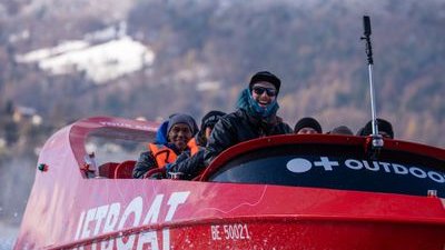 People on a red jet boat wearing jackets and hats, cruising on water with snowy mountains in the background.