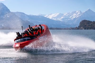 People in life jackets on a red jet boat speeding across a lake with snowy mountains in the background.
