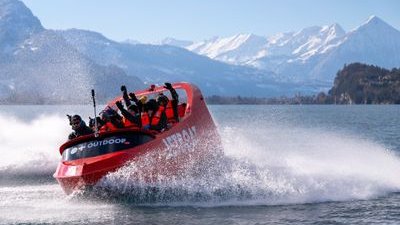 People in life jackets on a red jet boat speeding across a lake with snowy mountains in the background.