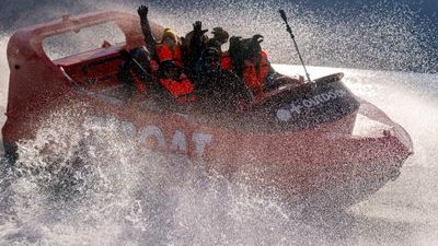People in life jackets on a red speedboat, splashing through water, with mountains in the background.