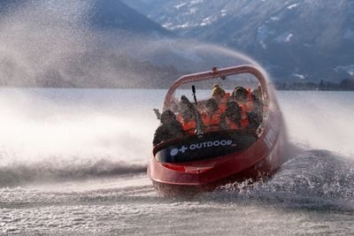 People in life jackets on a red speedboat, creating spray on a lake with mountains in the background.