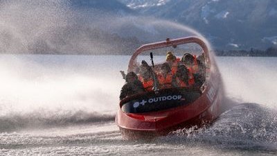 People in life jackets on a red speedboat, creating spray on a lake with mountains in the background.