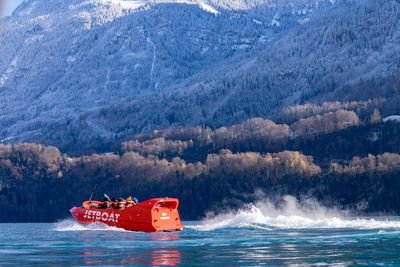 A red jet boat speeds across a lake with passengers in life jackets, set against snowy mountains.