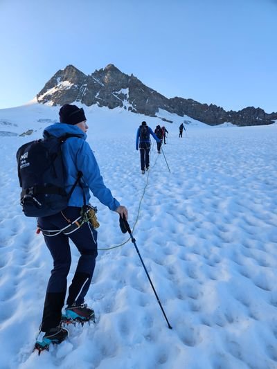 Several people with crampons, climbing harnesses, and hiking poles are ascending a snow-covered mountainside in an alpine setting.