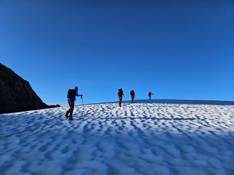 Four people with backpacks and hiking poles, some connected by rope, are hiking on a snow-covered mountain.