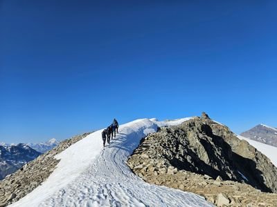 Several people with climbing gear and helmets are hiking in a line on a snow-covered mountain ridge in the Alps.