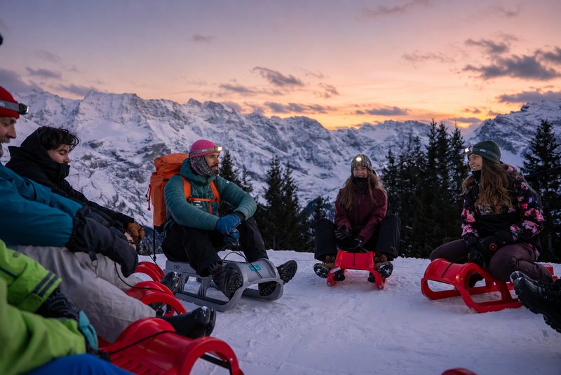 Group of people seated on red sleds in snow, wearing helmets and headlamps, backpacks, alpine mountains at sunset.