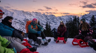 Group of people seated on red sleds in snow, wearing helmets and headlamps, backpacks, alpine mountains at sunset.