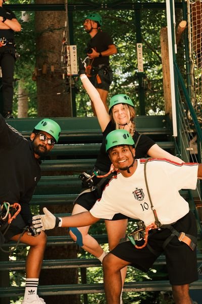 Drei Personen mit Kletterhelmen und Gurten beim Klettern an einer Metallstruktur im Wald.