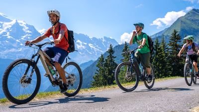 Drei Personen mit Helmen und Rucksäcken fahren Mountainbikes auf Bergstraße, Alpen im Hintergrund.