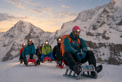 Gruppe auf Schlitten fährt im Schnee in alpiner Berglandschaft, trägt Helme, Stirnlampen, Rucksäcke