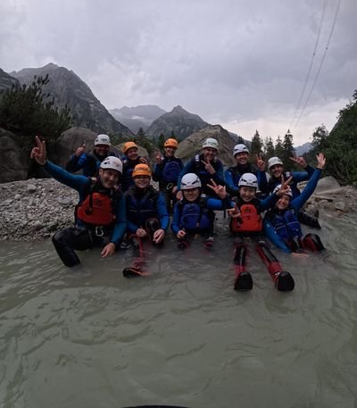 Gruppe von Personen mit Helmen, Neoprenanzügen und Schwimmwesten sitzt im Wasser vor Bergkulisse und Felsen.