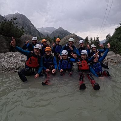 Gruppe von Personen mit Helmen, Neoprenanzügen und Schwimmwesten sitzt im Wasser vor Bergkulisse und Felsen.
