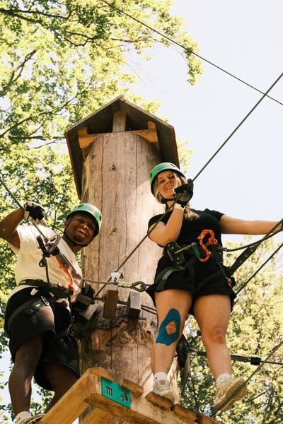 Zwei Personen mit Helmen und Klettergurten beim Seilrutschen in einem Wald mit Holzkonstruktion.
