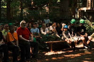 Gruppe von Personen sitzt im Wald auf Bänken, einige tragen Kletterhelme, im Hintergrund Kletterpark.