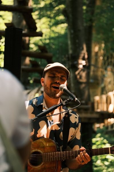 Person mit Hemd und Mütze spielt Gitarre und singt in natürlicher, bewaldeter Umgebung.