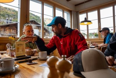 Drei Personen sitzen in einer Berghütte an einem Holztisch, eine Person trägt eine Kappe, keine sichtbare Sicherheitsausrüstung.