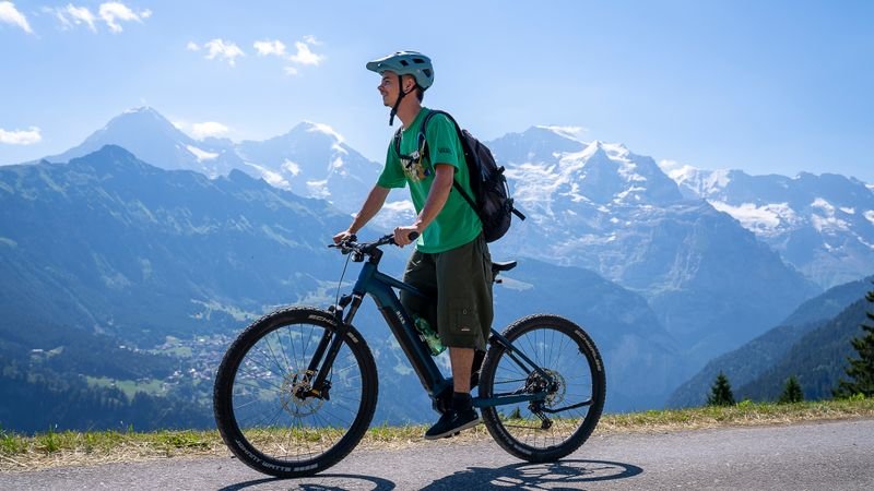 Person wearing a helmet and backpack riding an e-bike on a mountain road with snowy peaks in the background.