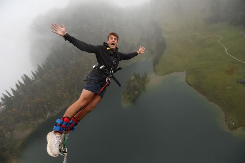 Person mit Sicherungsgurt und Fußfixierung beim Bungee-Jumping über einem See in bergiger Landschaft.