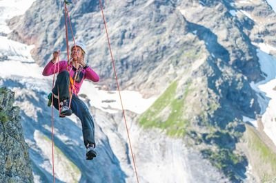 A woman is climbing up a rope using self-belay. In the background, the glacier landscape can be seen.