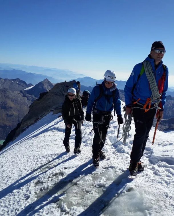 Three climbers with helmets and ropes ascend a snowy mountain ridge with a scenic view in the background.