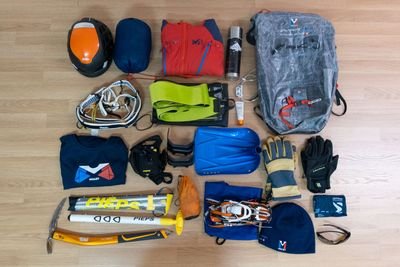 Diverses Bergsportmaterial für Gipfelskitouren ist am Boden ausgelegt. U.a. Helm, Pickel, Rucksack, Steigeisen, Schaufel, Sonde, LVS.