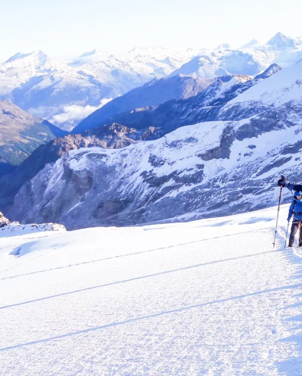Two people in helmets and harnesses trek on a snowy mountain with poles, surrounded by peaks.