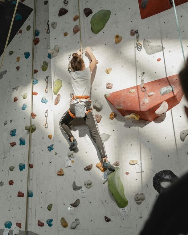 Person climbing on an indoor climbing wall with a harness, secured by another person.