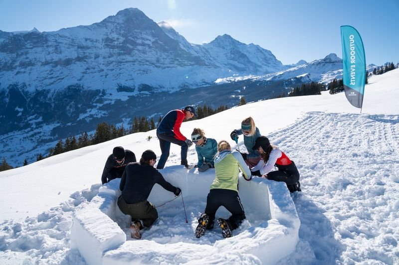 Personen bauen ein Iglu im Schnee, umgeben von Bergen. Einige tragen Winterkleidung und Mützen.