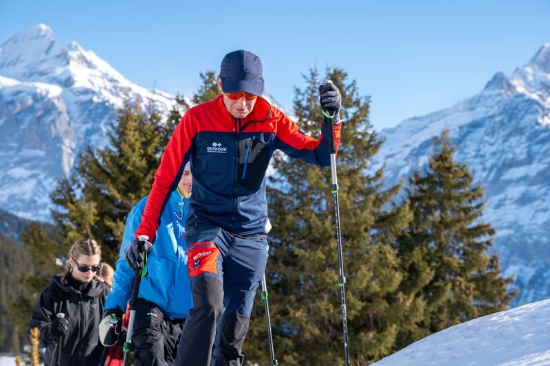 Personen mit Wanderstöcken und Winterkleidung gehen im Schnee vor Bergkulisse und Bäumen.