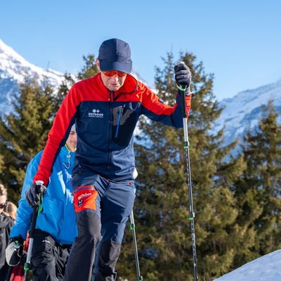 Personen mit Wanderstöcken und Winterkleidung gehen im Schnee vor Bergkulisse und Bäumen.