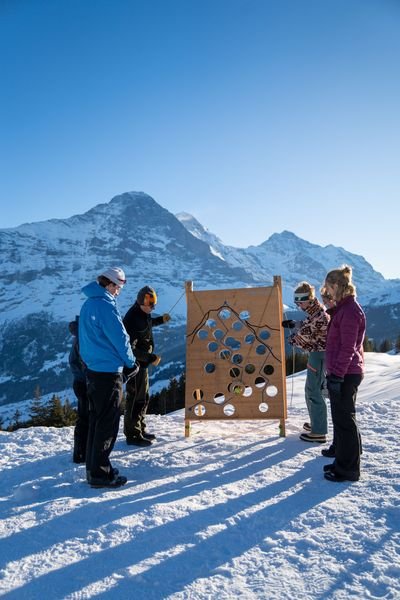 Personen stehen im Schnee vor einem Holzbrett mit Löchern, Berge im Hintergrund.