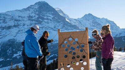 Personen stehen im Schnee vor einem Holzbrett mit Löchern, Berge im Hintergrund.