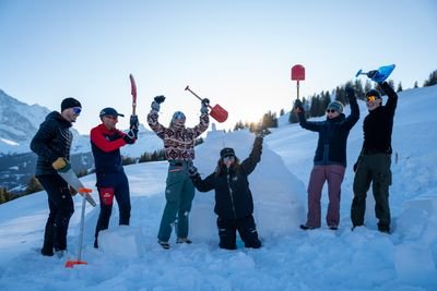 Sechs Personen mit Schaufeln im Schnee, umgeben von Bergen, tragen Winterkleidung und Sonnenbrillen.