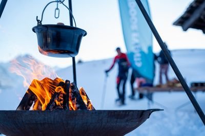 Ein Kessel hängt über einem Feuer im Schnee; im Hintergrund Personen mit Skistöcken und Banner.