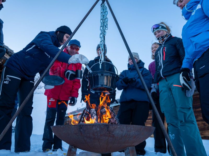 Eine Gruppe von Menschen steht im Schnee um ein Lagerfeuer mit einem hängenden Kessel.