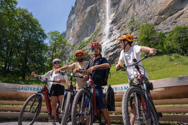 Vier Personen mit Helmen und E-Bikes vor Wasserfall und Felsen in bergiger, grüner Landschaft.