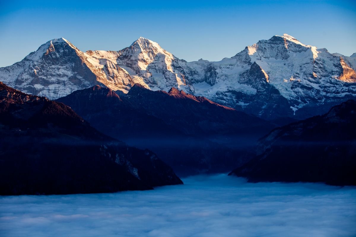 Blick auf schneebedeckte Alpenberge bei Sonnenaufgang, darunter Nebel im Tal.