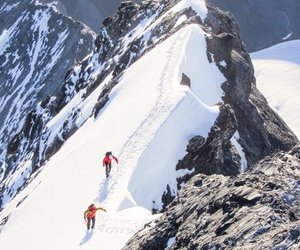 Two climbers in red jackets with backpacks and ice axes ascend a snowy mountain ridge.