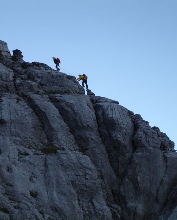 Two climbers in helmets and harnesses ascend a steep rocky mountain face, using ropes for safety.