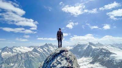 Person auf Berggipfel mit Kletterausrüstung und Seil, umgeben von verschneiten Alpen.