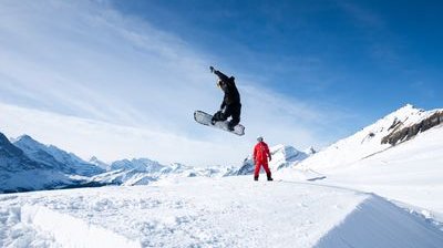 Snowboarder in mid-air jump on snowy slope, with another person in red suit watching, mountains in background.