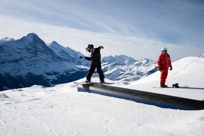 Snowboarder in schwarzer Ausrüstung auf einem Geländer, mit einem Führer in Rot in der Nähe, in einer verschneiten Berglandschaft.