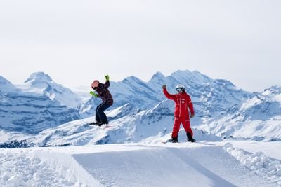 Snowboarder springt auf verschneiter Piste mit rotem Lehrer, im Hintergrund Berge.